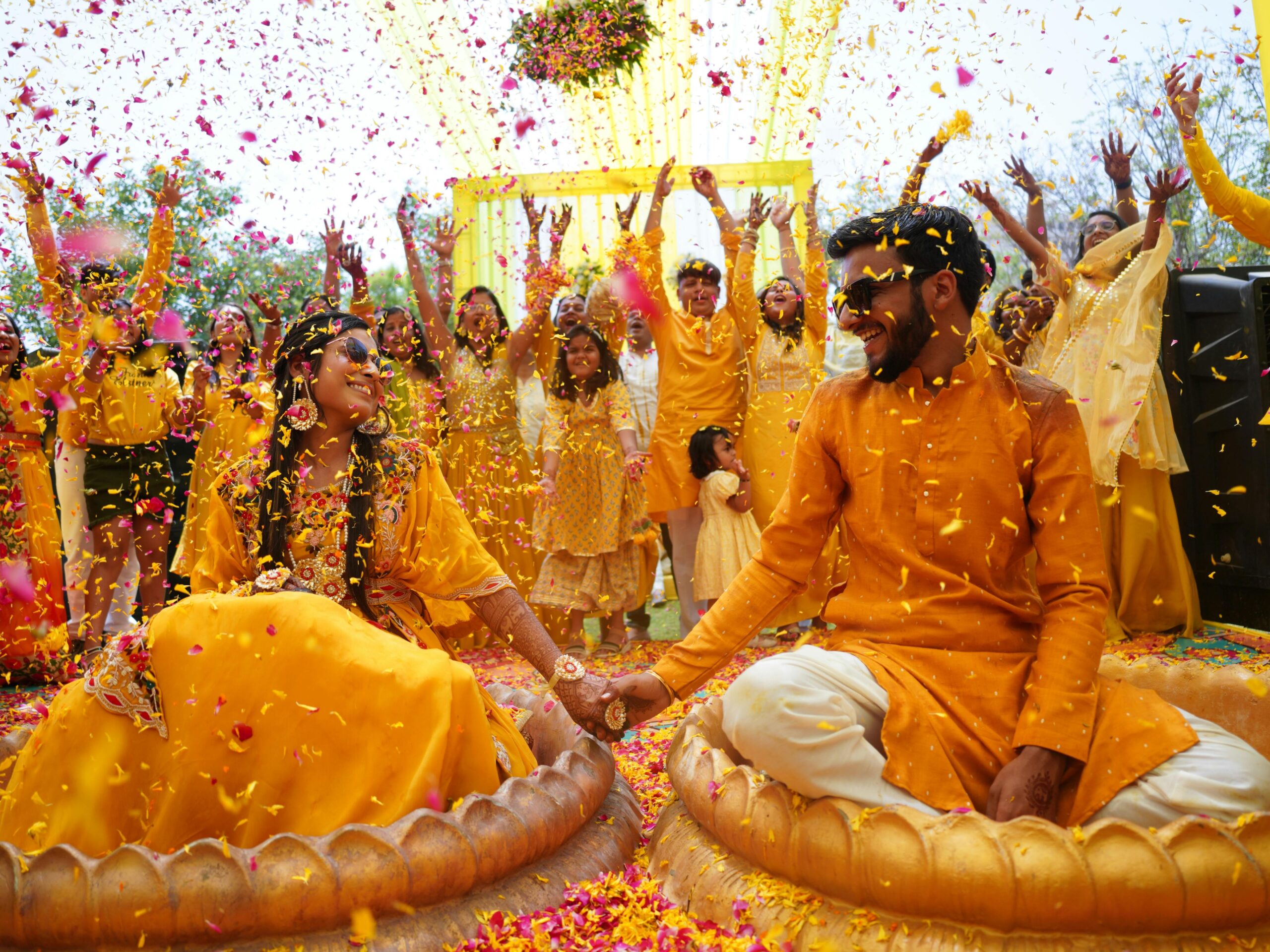 Joyful Indian haldi ceremony with a cheerful couple and guests throwing flower petals outdoors.